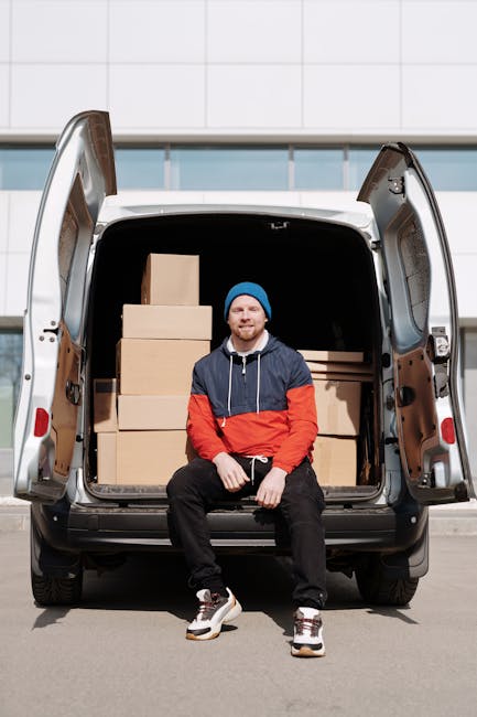 A man with a beard and glasses, dressed in a light blue shirt and dark trousers, is seated on a wooden chair outside a white delivery van parked on the street during daytime. The van's rear doors are 