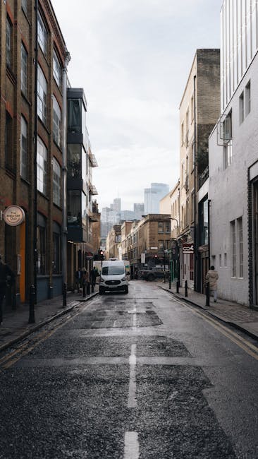 A narrow urban street scene featuring multi-story buildings on both sides, including a mix of brick and modern facades. The street surface appears wet, likely from recent rain, with visible patches of asphalt and a central dashed white line. Parked vehicles, including a white van prominently positioned in the middle of the street, are visible as part of the loading process. The van is positioned close to the pavement, with its rear doors possibly open, indicating ongoing furniture transport or home relocation activities. Several cardboard boxes and wrapped furniture items are seen on the pavement near the van or inside the vehicle, suggesting packing and moving preparations. In the background, taller buildings and cityscape elements are visible under a cloudy sky. The scene captures a typical street during a house removal operation, with no pedestrians or moving personnel visible, and subtle cues indicating the activities involved in packing, loading, and transporting household items, as associated with professional removals services such as those offered by Man with Van Enfield.