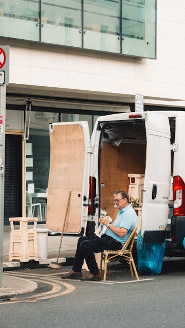 A man with glasses, wearing a light blue shirt and dark trousers, sits on a wooden chair outside a white moving van with its rear doors open, revealing a partially loaded interior containing cardboard boxes and packing materials. The van is parked on a street in front of a modern building with large glass windows and a beige facade. Several wooden crates and packing boards are arranged nearby on the pavement. The scene depicts a home relocation process involving furniture transportation and packing activities, with natural daylight illuminating the area. Manwithvanenfield.org specializes in removals and moving services, highlighting professional furniture transport and loading procedures in urban environments.