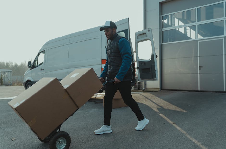 A man with a cap, dark vest, and casual clothing is engaged in the loading process for a home relocation, pushing a two-wheeled trolley with two large cardboard boxes wrapped in packing tape. He is outdoors, on a paved surface beside a white commercial van with its rear doors open, revealing a spacious interior. The background features a modern building with glass panels, and the scene is lit by natural daylight, indicating a clear weather condition. The cardboard boxes are of different sizes, with one slightly smaller and stacked on top of the larger one, suggesting organized packing for moving logistics. The man is walking towards the back of the van, part of a furniture transport and packing operation conducted by Man with Van Enfield, a company specialising in removals and office moves. This image captures the typical loading activity involved in professional removals, emphasizing careful handling of packed belongings and efficient transport preparation.