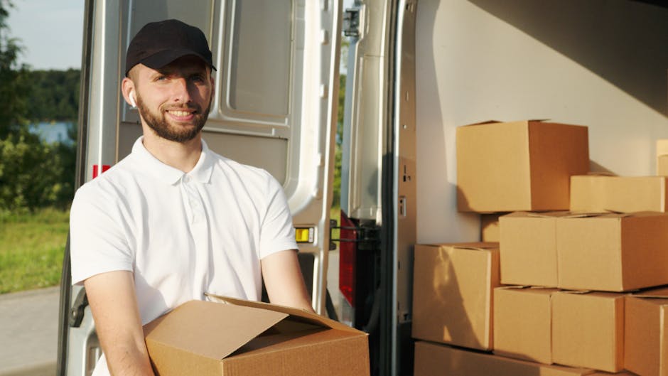 A man with a beard and wearing a black cap and white polo shirt is standing outside next to an open van, holding a cardboard box. The van's rear door is open, revealing several stacked cardboard boxes inside, some wrapped in plastic with packing materials visible around them. The scene is set outdoors on a sunny day with greenery and trees visible in the background. The man appears to be involved in a home relocation or furniture transport process, with the boxes positioned for loading or unloading as part of a house removal service. The environment suggests a professional moving operation, with the focus on careful handling of the boxes and organized packing materials, aligned with the services offered by Man with Van Enfield, supporting the logistics of house removals, packing, and furniture transport.