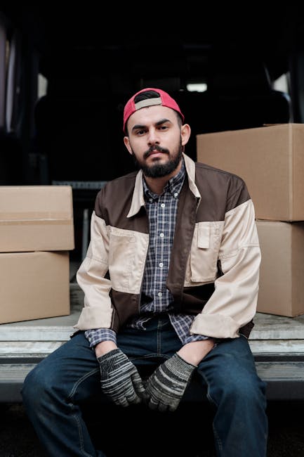 A man with a cap, dark vest, and casual clothing is engaged in the loading process for a home relocation, pushing a two-wheeled trolley with two large cardboard boxes wrapped in packing tape. He is outdoors, on a paved surface beside a white commercial van with its rear doors open, revealing a spacious interior. The background features a modern building with glass panels, and the scene is lit by natural daylight, indicating a clear weather condition. The cardboard boxes are of different sizes, with one slightly smaller and stacked on top of the larger one, suggesting organized packing for moving logistics. The man is walking towards the back of the van, part of a furniture transport and packing operation conducted by Man with Van Enfield, a company specialising in removals and office moves. This image captures the typical loading activity involved in professional removals, emphasizing careful handling of packed belongings and efficient transport preparation.