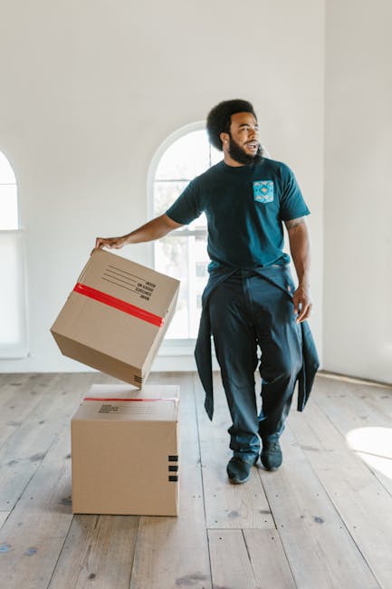 A man with a dark beard and curly hair, wearing a navy blue t-shirt with a logo on the chest and dark trousers, is inside an empty, well-lit room with white walls and wooden flooring. He is in the process of moving, holding a cardboard box with red packing tape in his right hand, preparing to place it on top of another box on the floor. Behind him is a large, arched window allowing natural light to fill the space. The scene captures a moment of furniture transport or packing during a house relocation, fitting with the services offered by Man with Van Enfield. The setting emphasizes the interior environment of a home undergoing packing or furniture transport as part of a professional move.