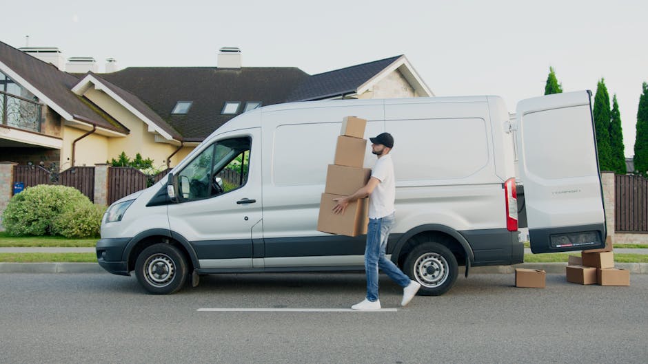 A man wearing casual clothes and a black cap is loading three large cardboard boxes into the back of a white panel van parked on a residential street. The van is positioned parallel to the curb, with its rear doors open, revealing the interior used for home relocation. Several additional cardboard boxes are placed on the pavement nearby, likely ready for transport. The background features a multi-storey house with a sloped roof, small windows, and a front garden enclosed by a wooden fence. Tall, narrow evergreen trees are visible behind the house, and the scene is illuminated by natural daylight, suggesting good weather. The overall setting depicts a typical logistics process of furniture transport and packing during a house move, supported by the presence of the van and packed boxes, with the company Man with Van Enfield providing a quick and efficient removals service in the area.