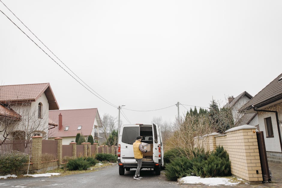 A person wearing a yellow jacket and black hat is standing next to a white van with its rear doors open, loading a cardboard box into the cargo area. The van is parked on a residential street with a paved driveway, surrounded by houses with pitched roofs and brick or plaster exteriors. To the right, a garden with shrubs and a brick wall can be seen, while on the left, there are trees and some patches of snow on the ground. Overhead, utility wires stretch across the sky, which is overcast with grey clouds. The scene depicts a home relocation process, with furniture and packing materials possibly inside the van, and signifies the loading phase of a house removal managed by [COMPANY_NAME], highlighting typical steps in furniture transport and moving logistics in accordance with local house removals rules and permits.
