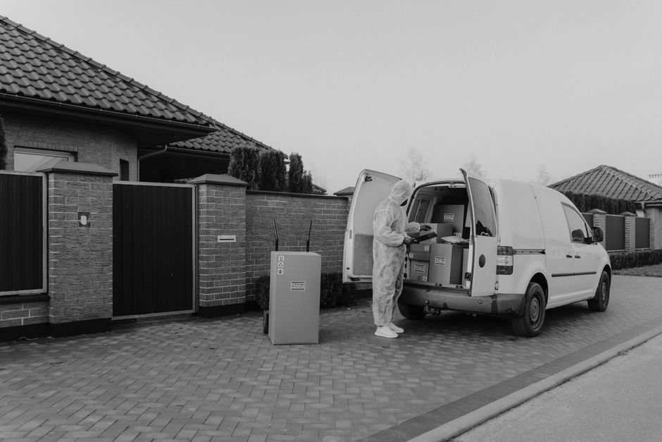 A person wearing a yellow jacket and black hat is standing next to a white van with its rear doors open, loading a cardboard box into the cargo area. The van is parked on a residential street with a paved driveway, surrounded by houses with pitched roofs and brick or plaster exteriors. To the right, a garden with shrubs and a brick wall can be seen, while on the left, there are trees and some patches of snow on the ground. Overhead, utility wires stretch across the sky, which is overcast with grey clouds. The scene depicts a home relocation process, with furniture and packing materials possibly inside the van, and signifies the loading phase of a house removal managed by [COMPANY_NAME], highlighting typical steps in furniture transport and moving logistics in accordance with local house removals rules and permits.